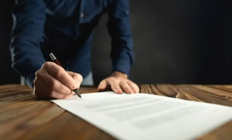 Mano firmando un documento con pluma sobre un escritorio de madera